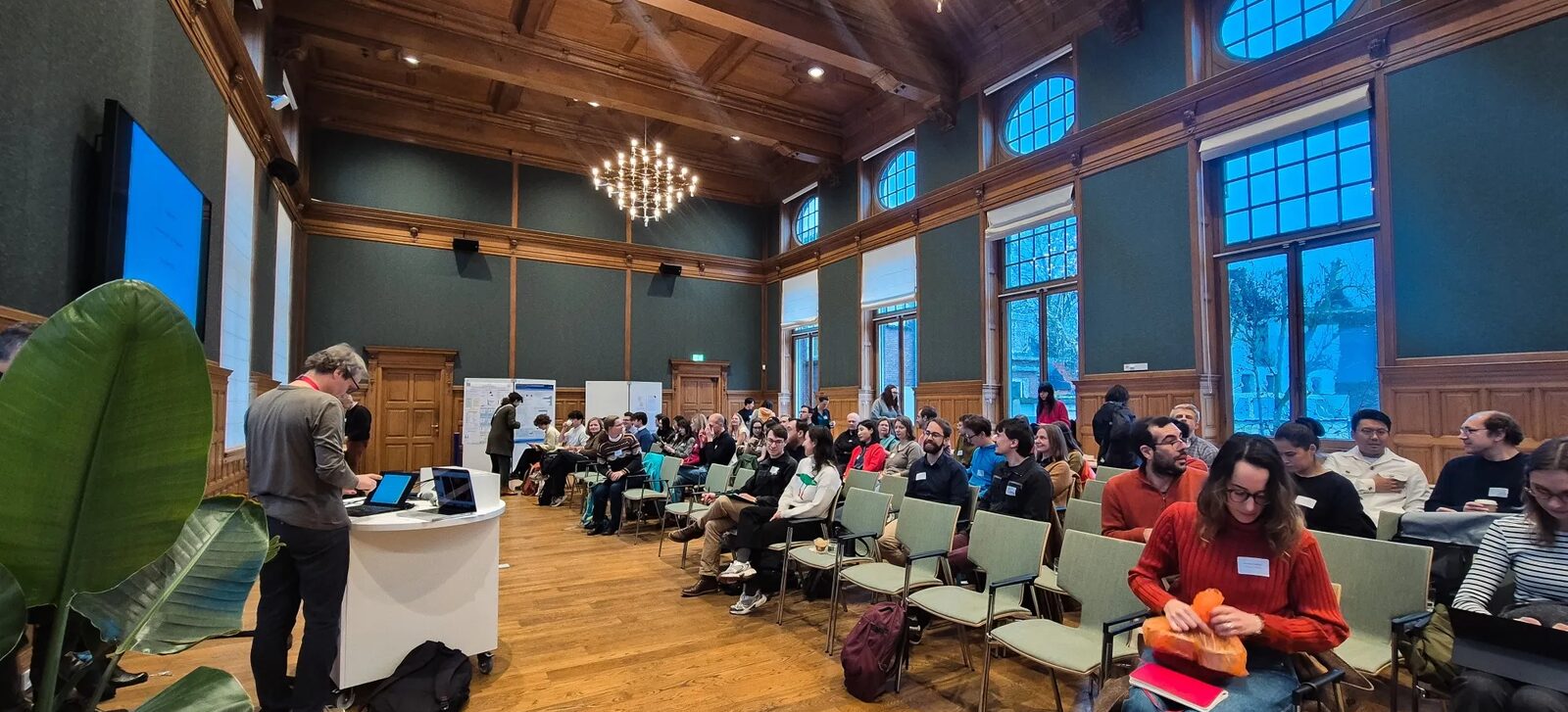 Audience seated in a wood-panelled hall with a chandelier, with a speaker at a lectern on the left