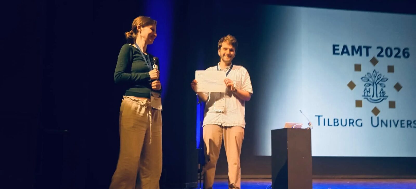 Two people on a stage, one holding a certificate, in front of a screen showing the EAMT 2026 and Tilburg University logos