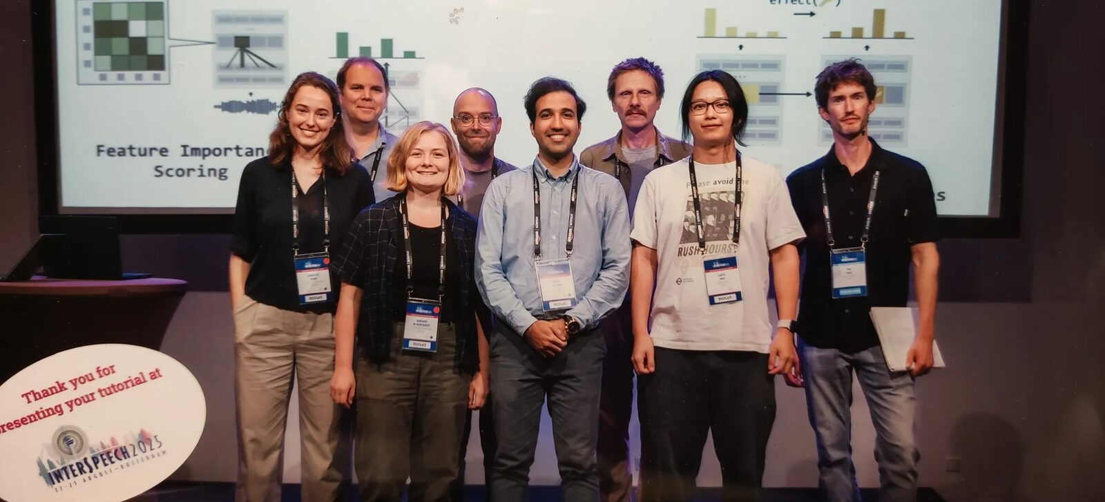 Eight presenters on a stage in front of a slide. Left to right: Charlotte Pouw, Willem Zuidema, Marianne de Heer Kloots, Martijn Bentum, Hosein Mohebbi, Grzegorz Chrupała, Gaofei Shen and Tom Lentz.